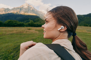 Naklejka premium Young woman with ponytail wearing wireless earbuds hiking in green meadow with mountain background enjoying nature and active lifestyle outdoors in fresh air.