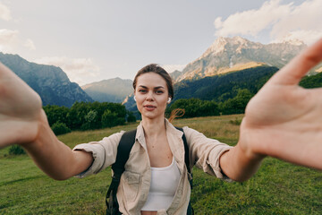 Naklejka premium A young woman in casual clothes takes a selfie outdoors in a green meadow with mountains in the background, enjoying nature and an active lifestyle.