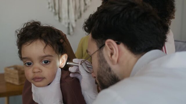 Close up shot of young male doctor checking ear of toddler patient with flashlight while sitting with African American mother during home visit