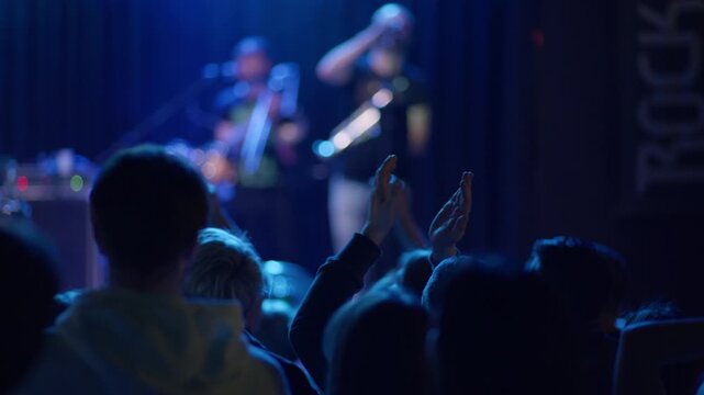 Fan claps raised hands at rock concert, blurred guitarist with guitar on stage behind, blue teal stage light, bokeh lights scattered in scene