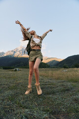 Naklejka premium Joyful woman jumping in a grassy field with mountains in the background, depicting outdoor lifestyle, freedom, and carefree adventure under a clear sky.
