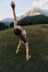 Naklejka premium A young woman dances in a grassy meadow, arms raised toward the sky, mountains in the background, conveying freedom, vitality, and a carefree outdoor lifestyle.