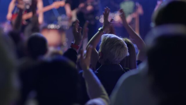 Young fan claps and raises hands from within crowd at live music event, band equipment and drum kit visible blurred in cool blue purple stage light behind