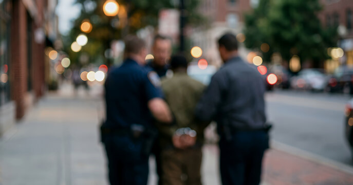 Out of focus image captures several officers holding a detained individual near parked cars, emphasizing anonymity, action, and a realistic portrayal of policing in a city environment.