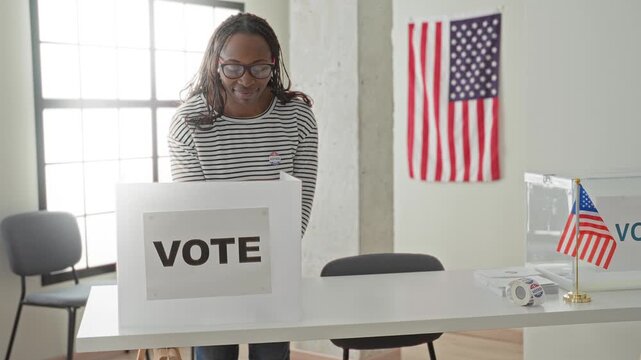 Woman at voting booth smiling and showing voted sticker beside transparent ballot box and american flag in a building; civic pride.