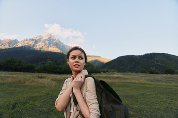 Naklejka premium A young woman with a backpack walks through a green meadow as distant mountains rise under a clear sky, symbolizing adventure and exploration.