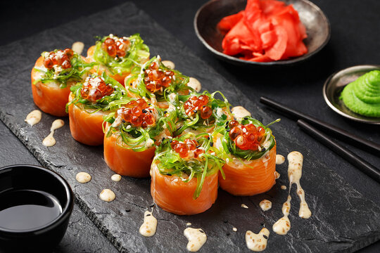 Sushi roll with salmon, wakame seaweed and peanut sauce garnished with red caviar and sesame seeds on black slate plate with soy sauce, wasabi and ginger over black concrete background. Close up view