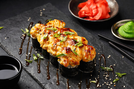 Baked warm sushi roll topped with chopped green onion and sesame seeds on black slate plate with soy sauce, wasabi and ginger over black concrete background. Close up view 