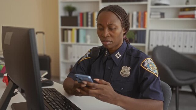 Young policewoman holding smartphone at desk inside building, looking down and tapping the screen with both hands; concentration duty responsibility.