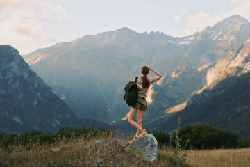 Naklejka premium A young woman with a backpack balances on a sun-warmed rock in a wide mountain meadow, gazing toward distant peaks with a sense of exploration, freedom, and awe.