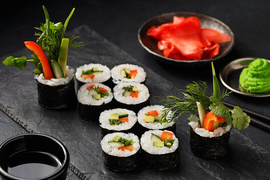 Veggie sushi roll with cucumber, avocado, bell pepper and greens on black slate plate with soy sauce, wasabi and ginger over black concrete background. Close up view