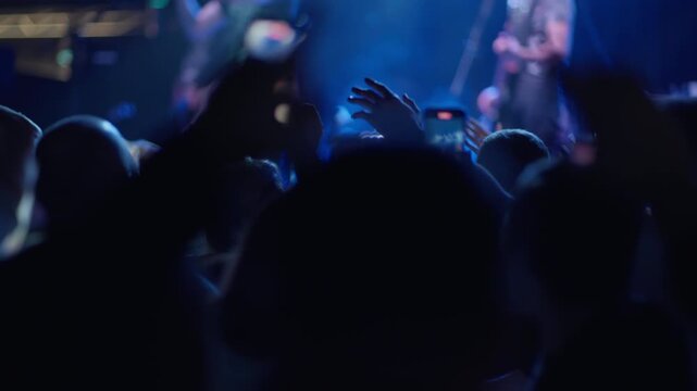 Several fans hold phones filming toward lit stage, performer visible blurred in background, blue stage atmosphere, venue wall visible at side