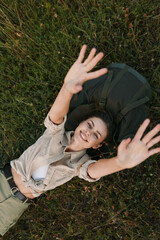 Naklejka premium A smiling woman relaxes on grass outdoors, arms raised in a carefree pose. Casual outfit, natural light, and green surroundings convey a joyful, laidback lifestyle.