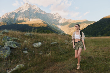 Naklejka premium A woman hiking in a sunlit field with towering mountains in the distance, capturing outdoors lifestyle, adventure, and nature within a vivid landscape for stock use.