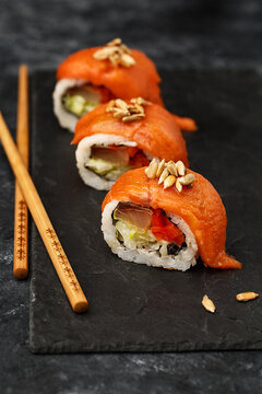 Three pieces of modern sushi roll with salmon, yellowtail, bell pepper and romano lettuce, decorated with fried sunflower seeds on black slate plate with chopsticks. Side view, close up