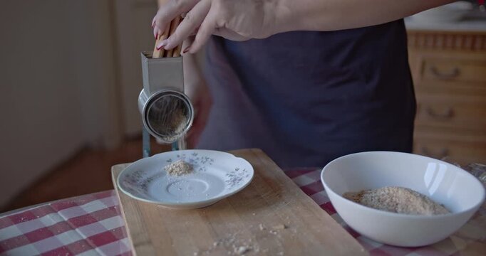 A woman uses a manual grinder to crush biscuits into fine crumbs, preparing a key ingredient for a delicious homemade dessert in her kitchen.