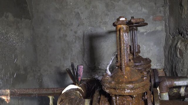Static shot of water leak from heavily corroded gate valve and flange on metal pipes in damp concrete basement with makeshift sealing tape repair attempts