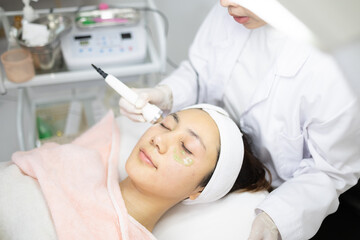 Naklejka premium Woman relaxes during facial treatment. Professional uses device gently on her face. Her eyes are closed, face calm and peaceful. Clinic environment appears clean and modern. Ideal for beauty, skincare