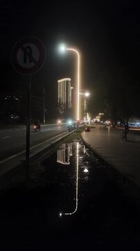 4K night street: Bright LED light reflecting in a puddle, featuring a No U-turn sign, heavy traffic, and a child on a tricycle with a luxury hotel skyscraper in the background. Urban safety scene.