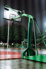 Green Basketball Hoop on Wet Outdoor Court at Night with Trees and Lights in Background © chuangxin