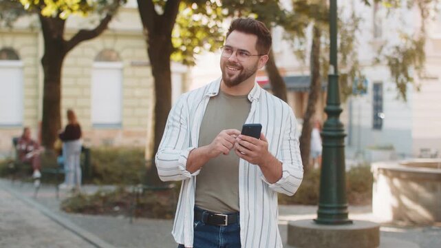 Happy successful satisfied Caucasian man young guy male traveler mobile phone user holding smartphone texting chatting social media tourist online GPS map outdoor urban city street. Town lifestyles.
