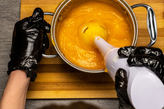 Top view chef in black gloves uses immersion blender to grind and emulsify pumpkin in saucepan in hot state. Cooking pumpkin soup for vegetarian. Vegetable dietary proper diet containing iron and zinc