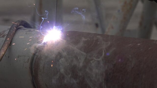 close-up of a gloved worker arc-welding a circumferential joint on a large-diameter metal pipe. The stick electrode strikes the seam and ignites a brilliant white-hot arc