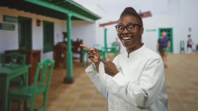 Woman chef points finger and smiles, wearing chef coat and glasses in building courtyard with green chairs and table; joy hospitality warmth.