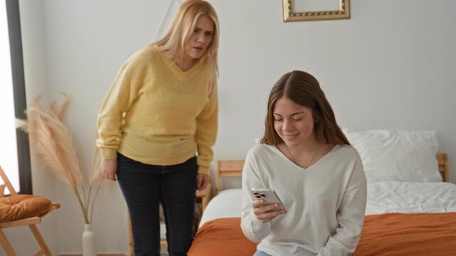 Woman reaches across and takes smartphone from seated daughter on bed in building while pointing and checking the screen; tension conflict.