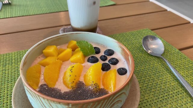 Healthy smoothie bowl topped with fresh mango, orange slices, blueberries and chia seeds, served with a creamy iced cappuccino on a wooden table. Bright, nutritious breakfast in a cafe setting