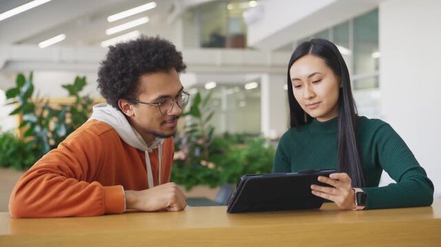 Engaged colleagues discussing something about what they watched on tablet screen. Male smiling. Asian woman explaining details with focused expression. Completing task together in office.