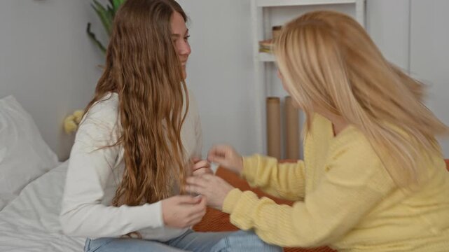 Mother and daughter laughing, hands playfully touching on a bed in a bedroom with pillows and a plant, blonde woman leaning over long haired teen; family bonding joy.