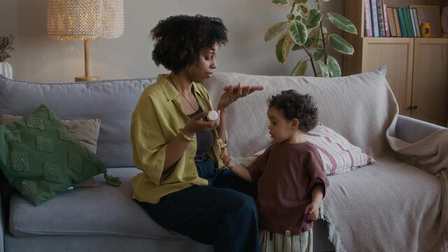 Medium long shot of African American woman holding medication and talking with doctor via smartphone while her little son standing by her in living room