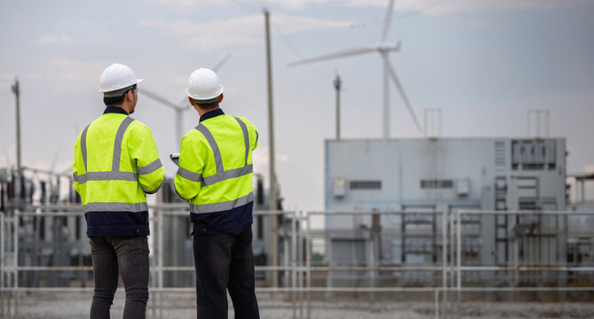 Two professional engineers analyzing renewable energy production at a wind turbine farm power plant substation. High-quality technical inspection and maintenance concept for sustainable electricity.