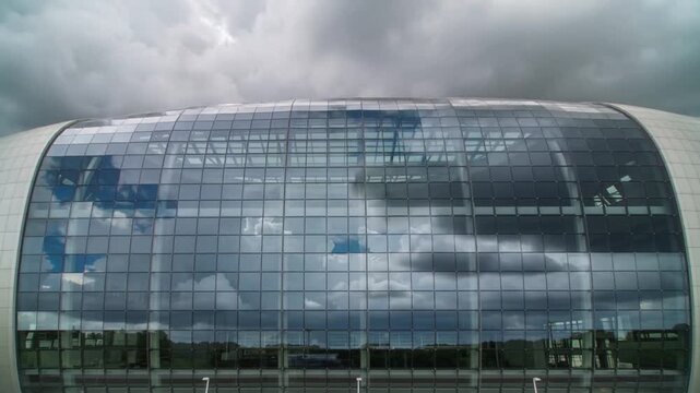 Showing curved glass facade at exterior reflecting sky with clouds shifting, revealing roof trusses