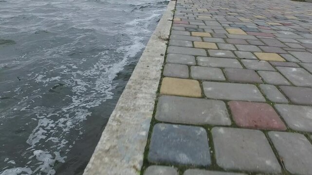 Low angle footage of seaside promenade with textured cobblestone pavement and waves along urban waterfront during cloudy weather