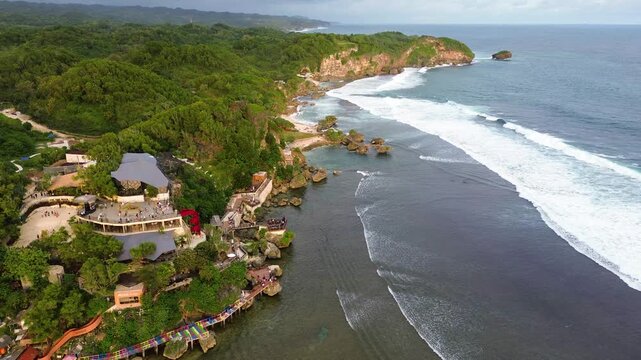 Aerial drone view of Drini Beach in Yogyakarta, Indonesia. Scenic tropical coastline with white sandy beach, clear turquoise water, small offshore island, and gentle ocean waves.