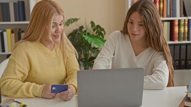Two women mother and daughter holding a blue creditcard beside an open laptop, hugging and smiling while seated in a home building; family celebration joy.
