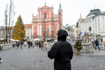 Fototapeta premium Rear view of man walking across Triple Bridge in Ljubljana Slovenia with Franciscan Church and Christmas tree in background capturing winter travel lifestyle atmosphere