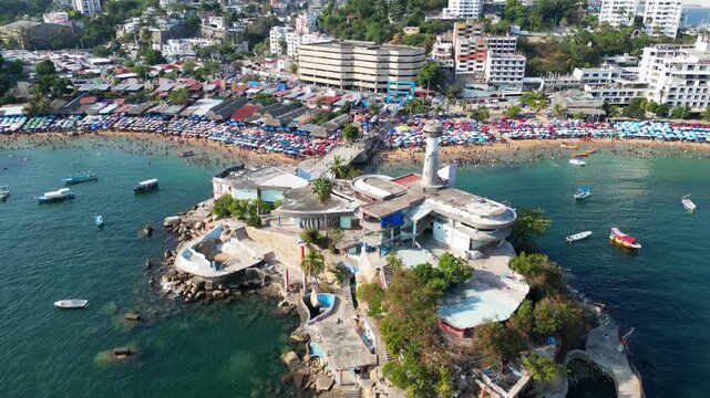 Vibrant aerial showing Caleta beach, Acapulco, Mexico. Crowds, colorful umbrellas, boats in bay. Popular summer vacation destination.