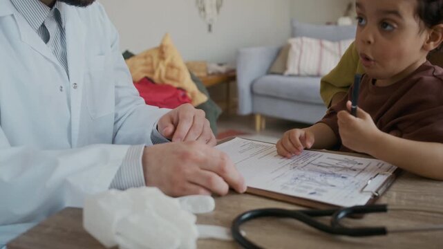 Medium close up handheld shot of small African American boy drawing on medical document while sitting at table with mother and male doctor during home visit