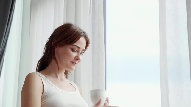 Happy caucasian woman open windows and drinking coffee in morning in bedroom at home, enjoy beverage with cozy for wellness, beautiful woman relax with beverage, self care, relaxation and lifestyle.