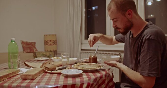 A young man intently prepares a sweet crepe at his dining table, adding chocolate spread and a sprinkle of sugar for a delightful, homemade meal.