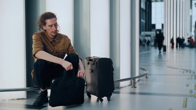 A young guy with long, curly hair sits on a metal or glass railing in a modern airport terminal, working on his laptop while waiting for a flight. 