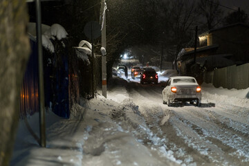 Obraz premium Snow-covered street scene at night with vehicles moving through deep snow and warm lights from houses visible in the background