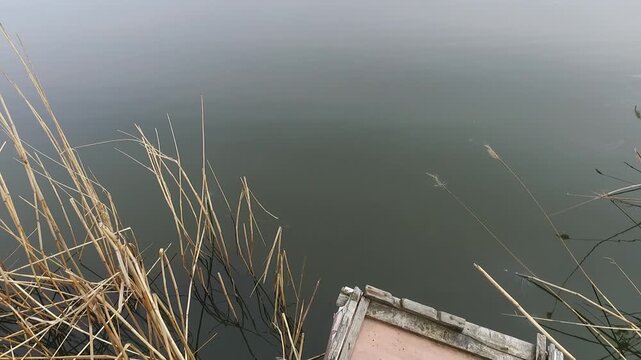 High-angle view of dry reeds and an old abandoned wooden fishing platform on a still overcast lake with soft water reflections in late winter