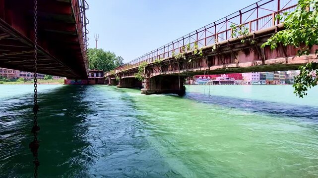 The swift, emerald-green currents of the Ganges flow between the weathered undercarriages of two parallel iron bridges in Haridwar, with colorful buildings and a distant Shiva Trishul monument.