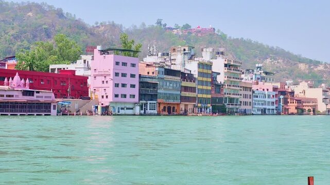 A variety of colorful ashrams and hotels, including a prominent pink structure and a deep red temple, line the bank of the turquoise Ganges river in Haridwar, with a hillside temple.