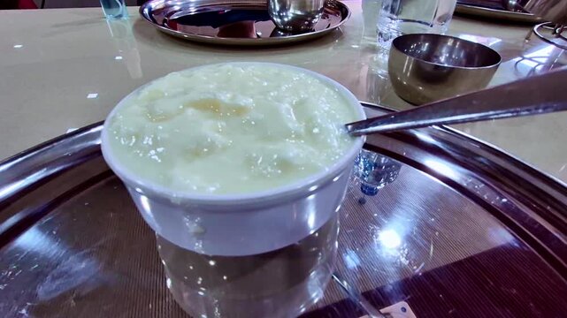 A bowl topped with thick, frothy curd sits on a shiny steel tray, surrounded by metal bowls, a water glass, and a hand resting near a smartphone on the table.