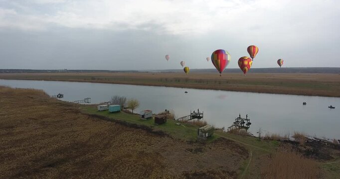Aerial view of colorful hot air balloon festival over wide flat river with golden reeds and steppe landscape. Fishing base with wooden piers and boats below. Overcast autumn sky. Multiple balloons in 